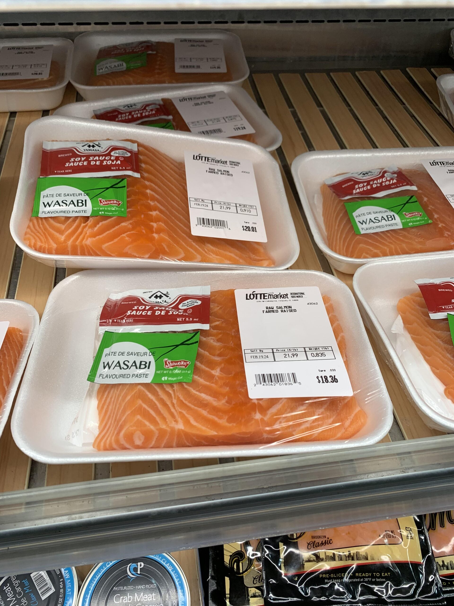 Packages of fresh salmon fillets in white foam trays, wrapped in plastic, with green wasabi and red sauce packets inside, displayed on a refrigerated grocery store shelf.