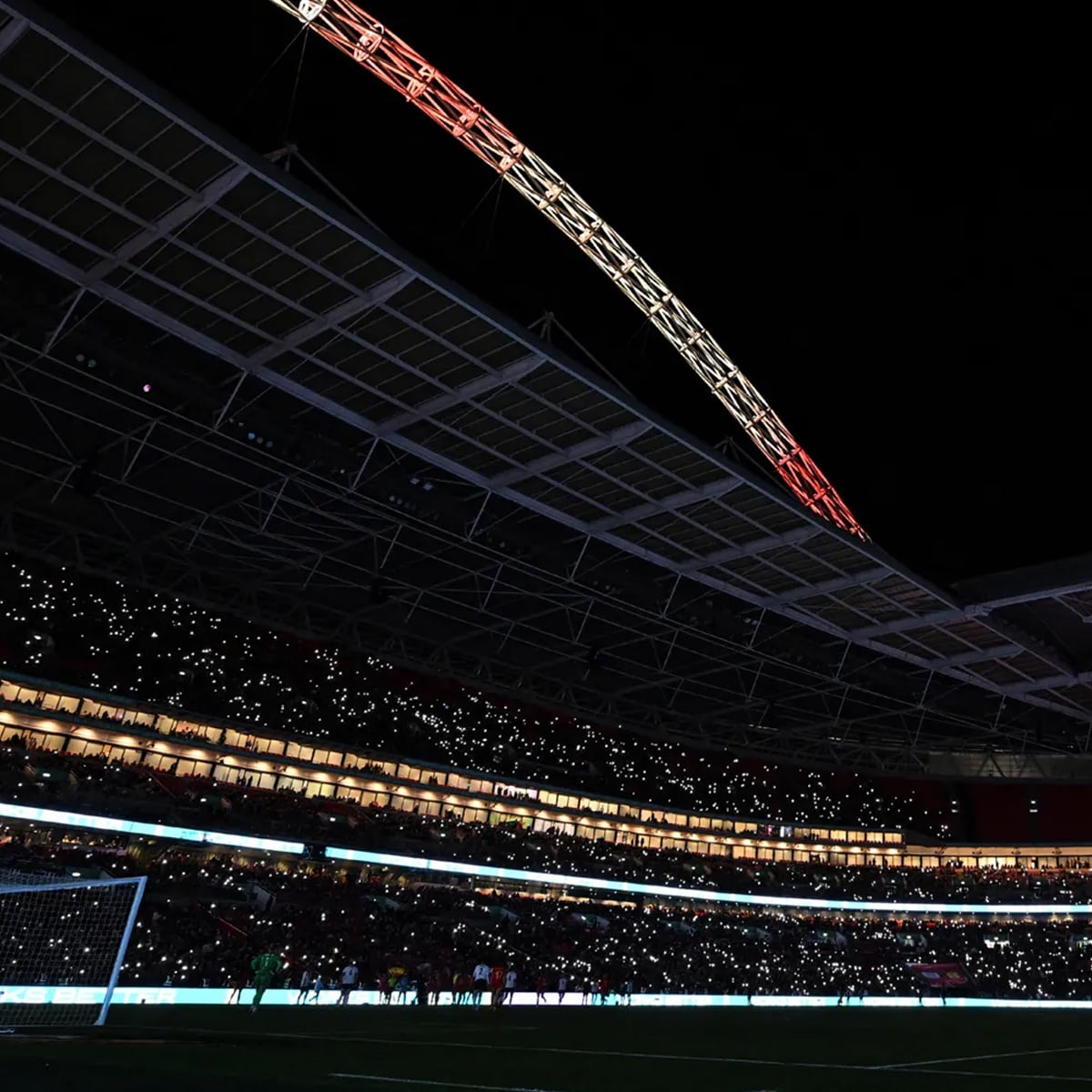 A night view of a stadium filled with spectators, many holding small lights. Part of a large illuminated arch is visible above the roof, and a soccer goal post is seen on the darkened field below.