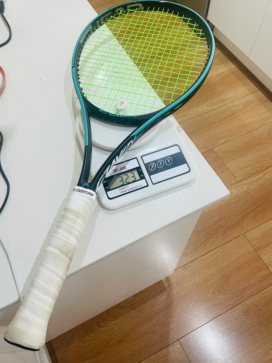 A tennis racket with a white overgrip rests on a digital scale, displaying a weight of 327 grams. The scale is on a white countertop, and the floor has a wooden finish.
