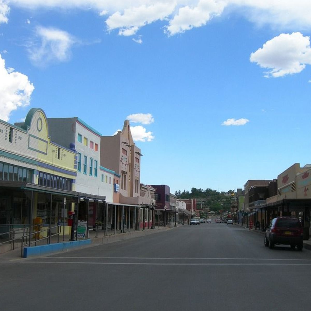A quiet small town street lined with colorful historic buildings under a bright blue sky with scattered clouds. There are a few parked cars but no people visible on the sidewalk or street.