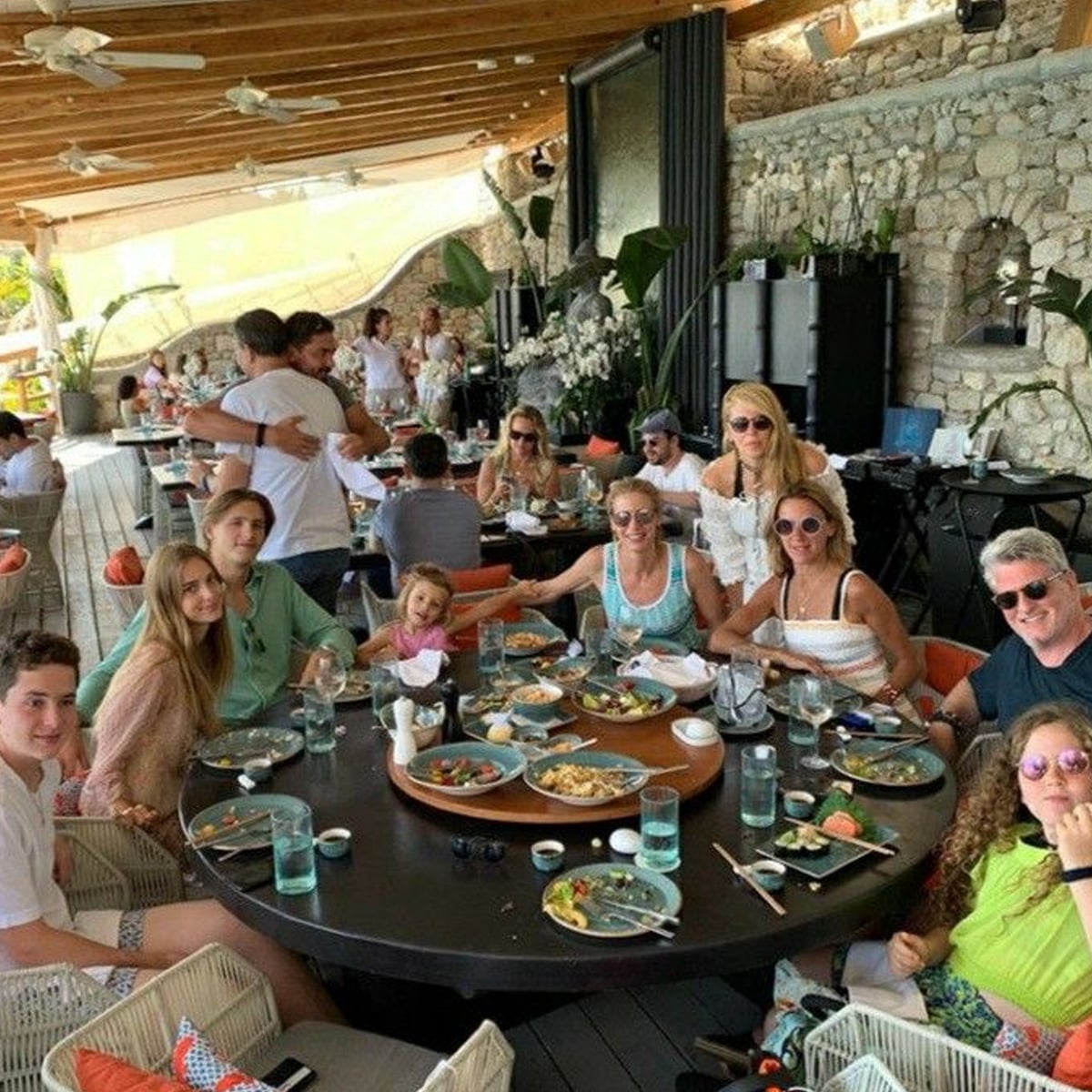 A group of people sit around a large restaurant table, enjoying a meal together. Plates of food and drinks are on the table, and the setting is bright with natural light and stone walls in the background.