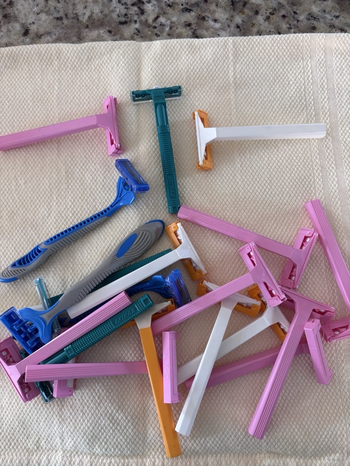 A pile of assorted disposable razors in pink, blue, white, and orange colors lies on a cream-colored textured towel on a countertop.