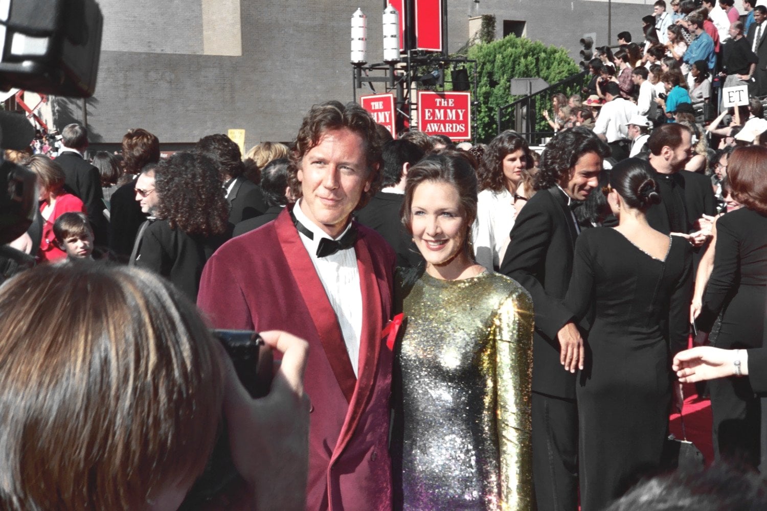 A man in a red velvet suit and a woman in a shiny gold dress pose together on a crowded red carpet at an awards event, with the Emmy Awards sign visible in the background.