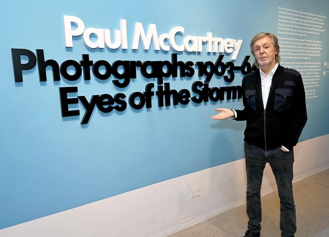 Paul McCartney stands beside a blue wall displaying large text: "Paul McCartney Photographs 1963-64 Eyes of the Storm," promoting his photography exhibit. He gestures toward the sign with one hand.