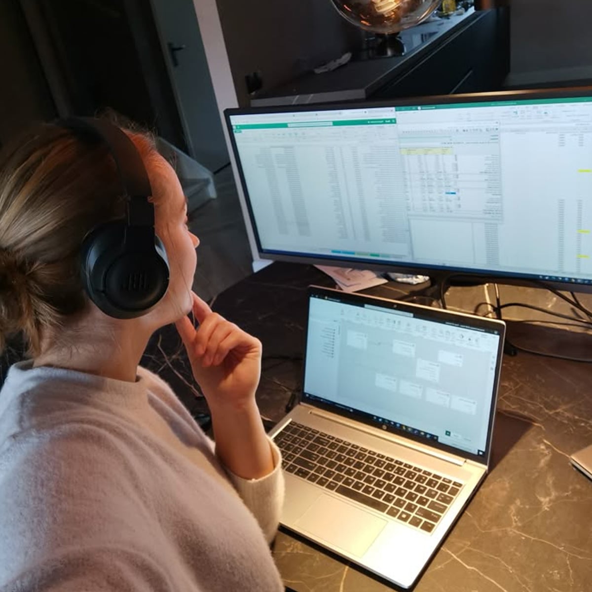 A person wearing headphones sits at a desk, looking at spreadsheets on two large monitors and a laptop, appearing focused while working from home.