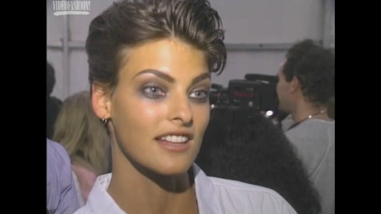 A woman with short brown hair and bold eye makeup smiles while speaking. She wears a white top and hoop earrings, and there are blurred people and equipment in the background.