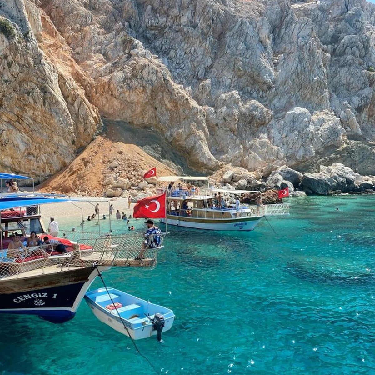 Turquoise water with two boats displaying Turkish flags near a rocky coastline; people are swimming and relaxing on the boats and small beach.