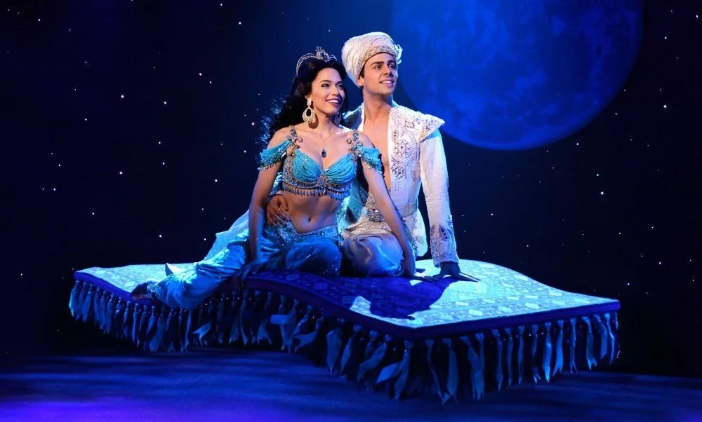 Two people dressed as Aladdin and Jasmine sit together on a flying carpet, surrounded by a starry night sky and a large blue moon, evoking the iconic magic carpet ride scene.