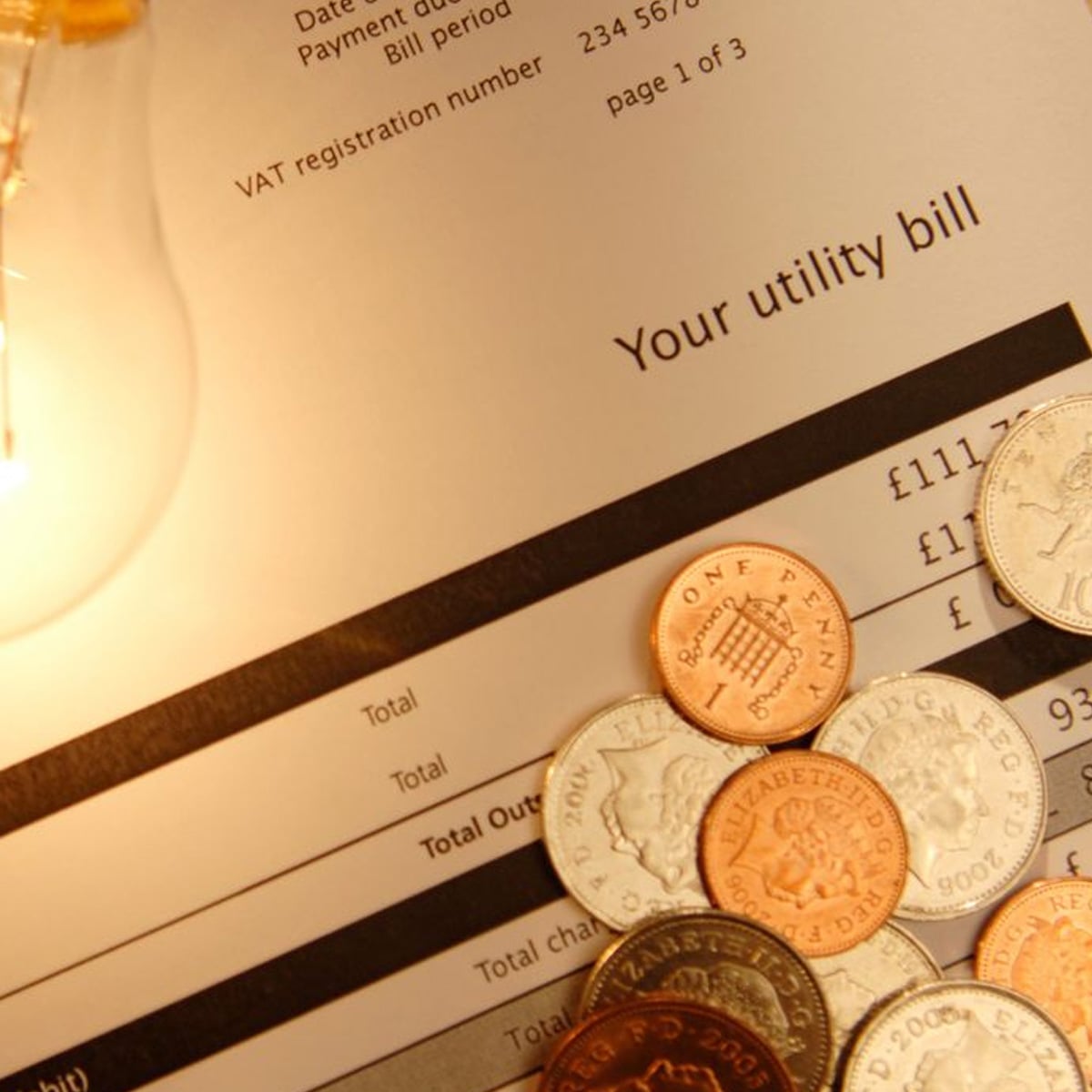 A close-up of a utility bill partially covered by various British coins, with a lit light bulb on the left side, symbolizing energy costs and household expenses.