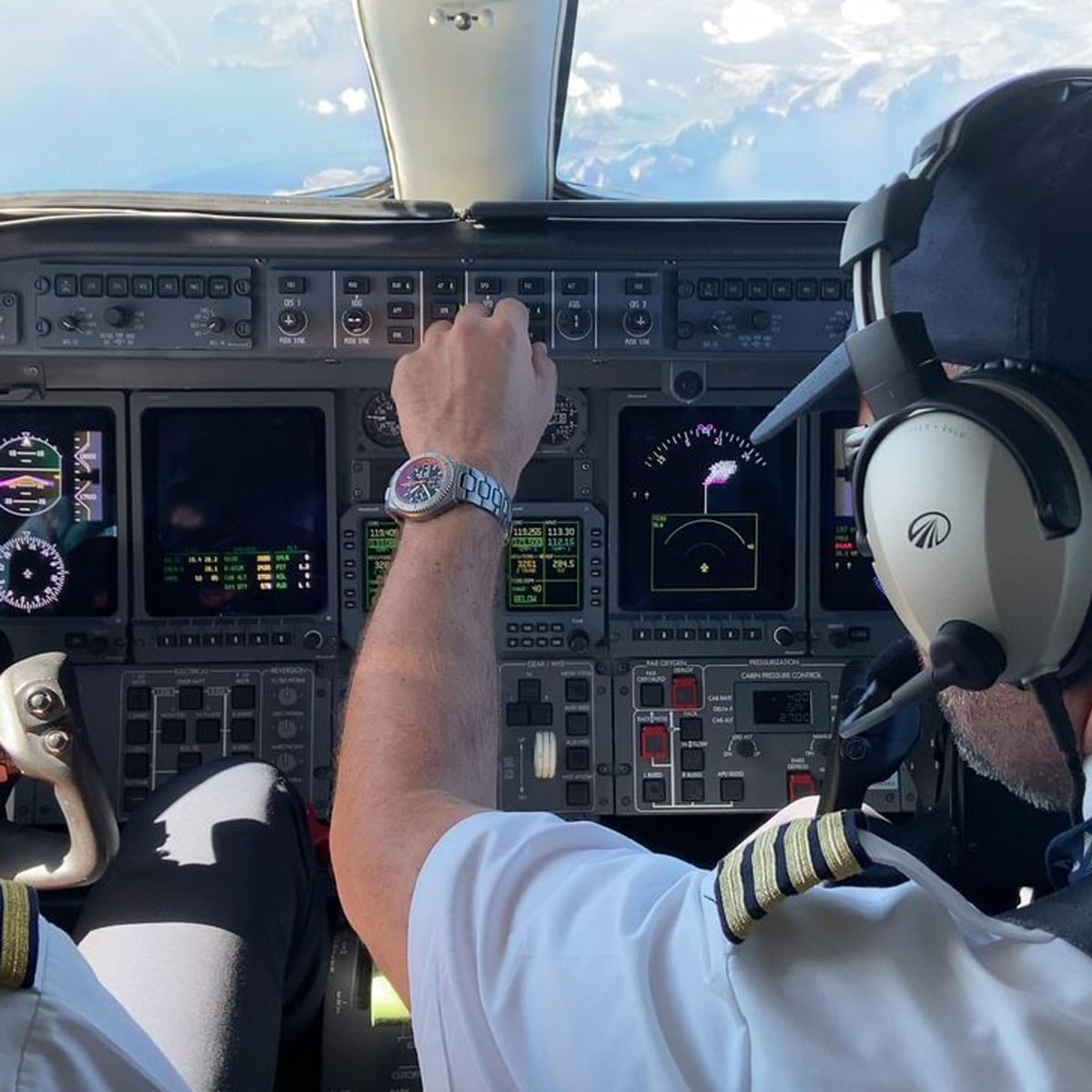 Two pilots in uniform sit in a cockpit, one adjusting controls on the instrument panel. Multiple screens display flight data, and both wear headsets. The view outside shows blue sky and clouds.