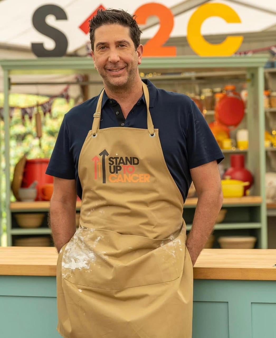 A man wearing a tan "Stand Up To Cancer" apron and a navy polo shirt stands in a kitchen setting with colorful utensils and large "S", "U", and "C" letters in the background.
