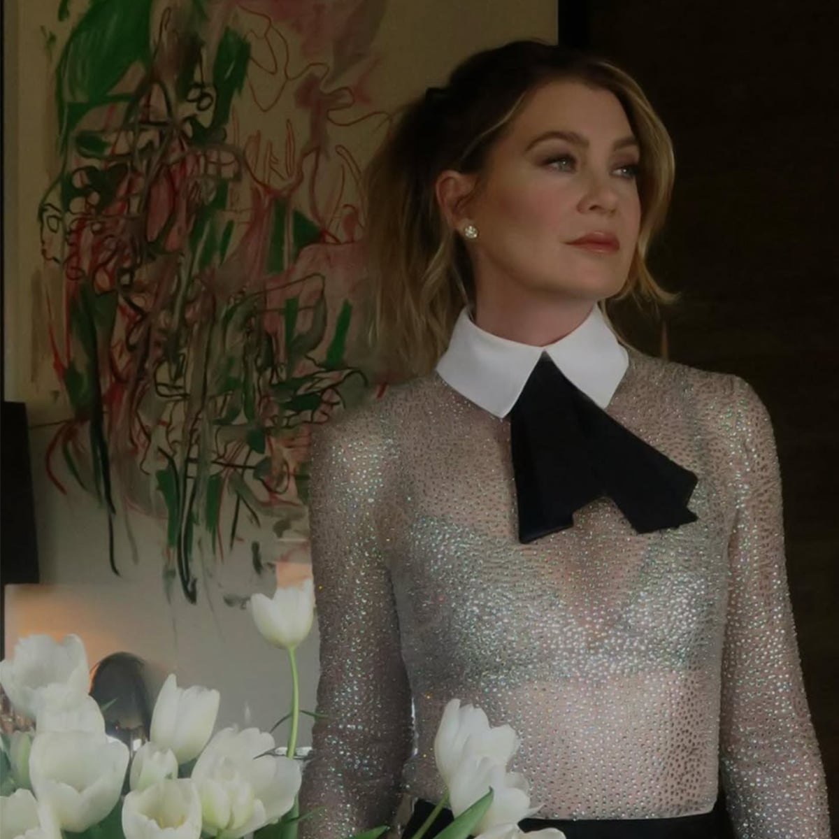 A woman wearing a sparkly, sheer top with a white collar and black bow stands indoors beside a vase of white tulips, with abstract art on the wall behind her.