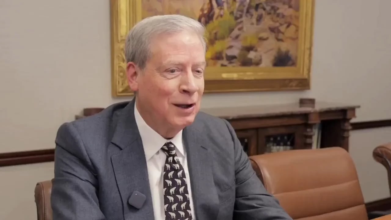 An older man in a gray suit and patterned tie sits at a table, speaking and smiling. There is a painting on the wall and a wooden cabinet in the background.