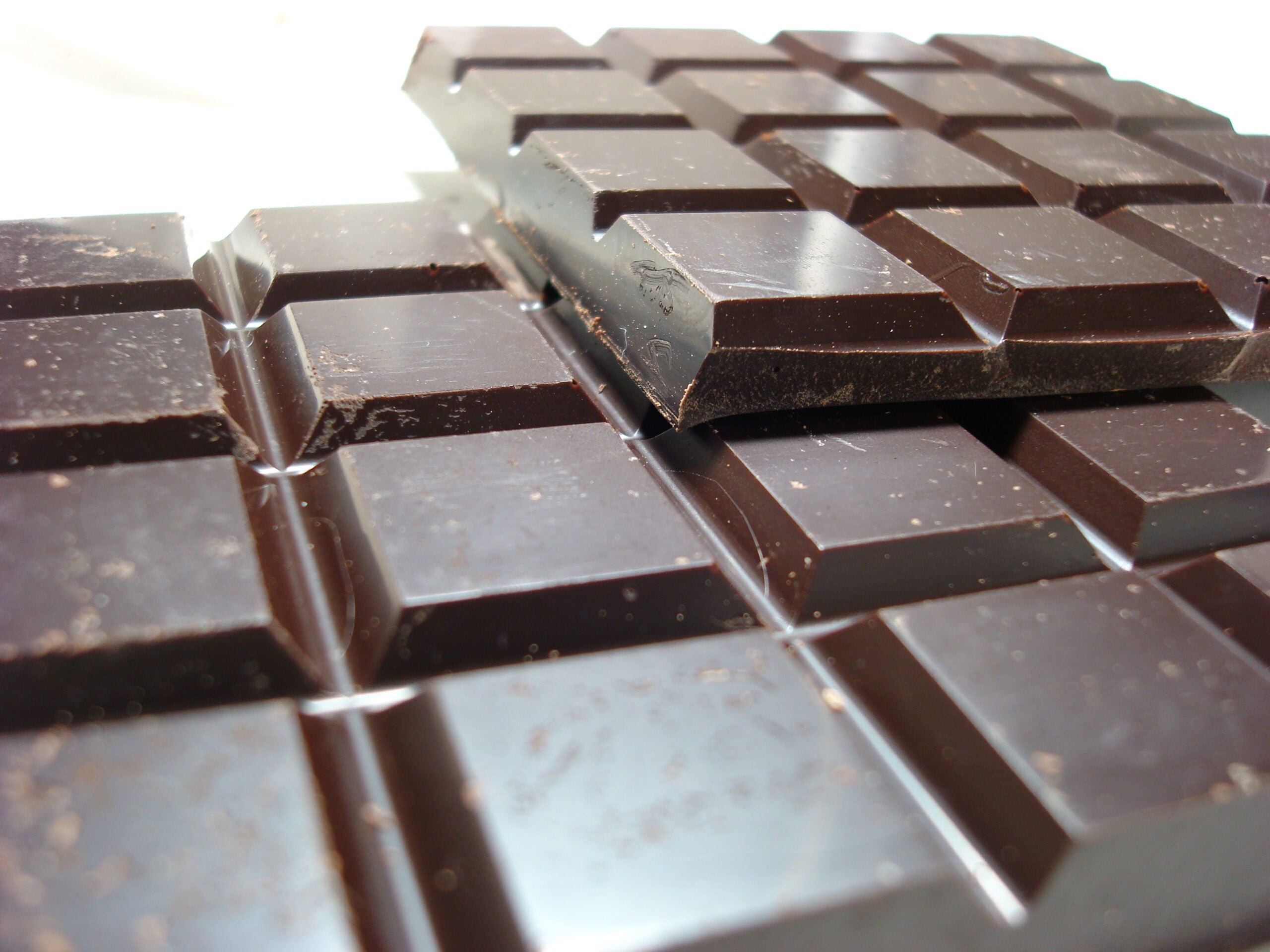 Close-up of several dark chocolate bars with a grid pattern, stacked on top of each other. The chocolate has a glossy surface with small bits of chocolate dust scattered across it.