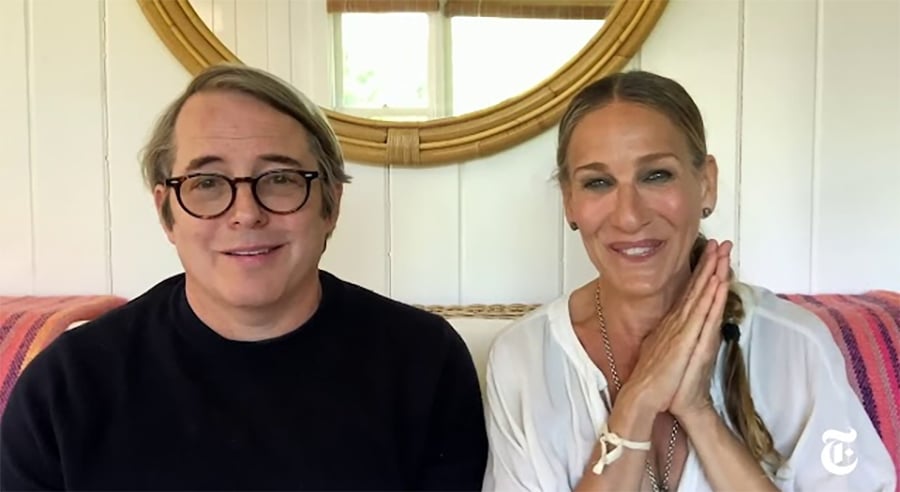 A man in glasses and a black shirt sits next to a smiling woman with braided hair, wearing a white blouse, in front of a round mirror and white paneled wall. Both are seated on a couch with a striped blanket.