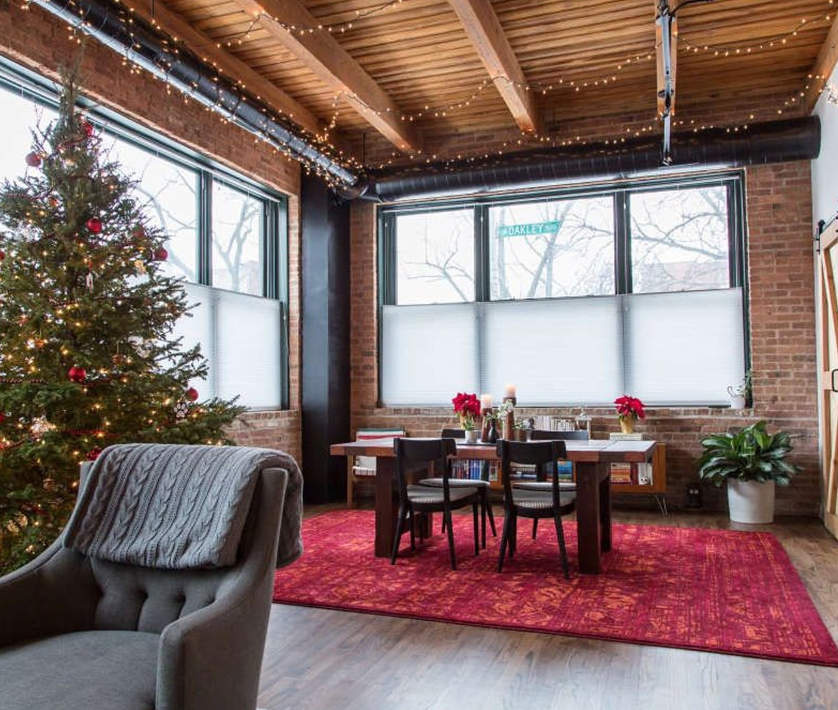 A cozy, modern dining area with large windows, a red rug, a decorated Christmas tree, wooden ceiling, exposed brick walls, and a gray armchair with a blanket. String lights add warmth to the festive space.