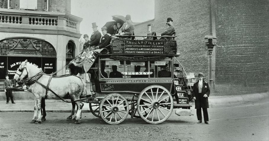 A black-and-white photo of a horse-drawn double-decker omnibus with passengers seated on top and inside. Two horses are harnessed to the carriage, and a man stands nearby on a cobblestone street in front of old buildings.