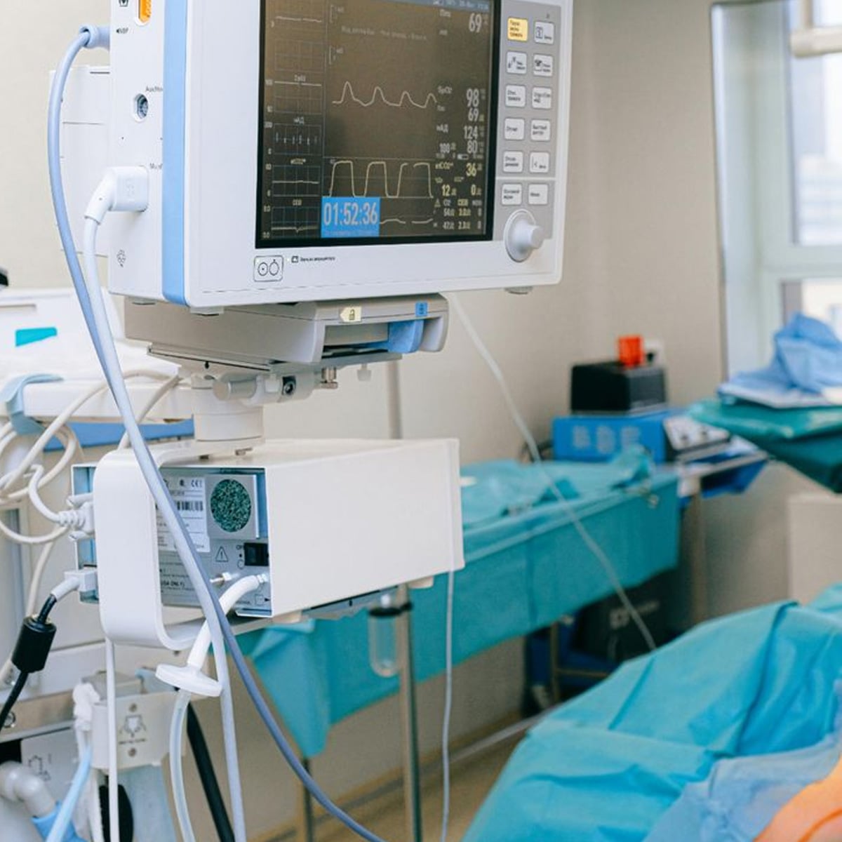 A medical monitor displays vital signs and a timer in an operating room. Tables with surgical drapes are visible in the background.