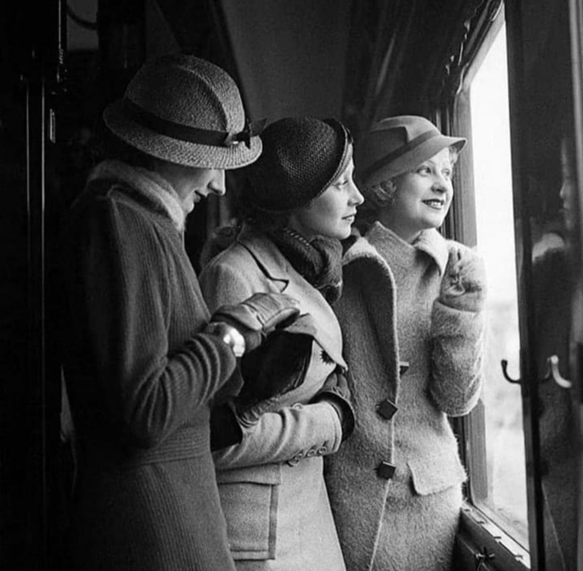 Three women in vintage coats and hats stand by a window. One reads a book or letter, the others look outside, smiling. The scene has a classic, nostalgic black-and-white style.