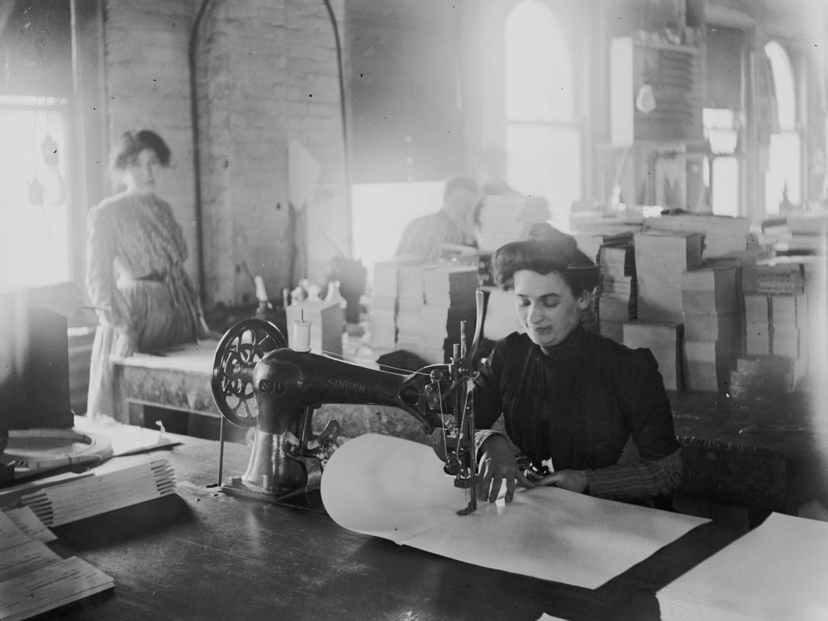 A woman uses a sewing machine to stitch papers at a worktable in an old factory, while others work in the background near large windows letting in daylight. Stacks of paper and documents are visible around the room.