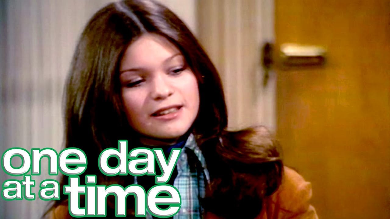 A young woman with long brown hair is indoors, looking down and smiling slightly. The words "one day at a time" are written in bold white and green text in the lower left corner.