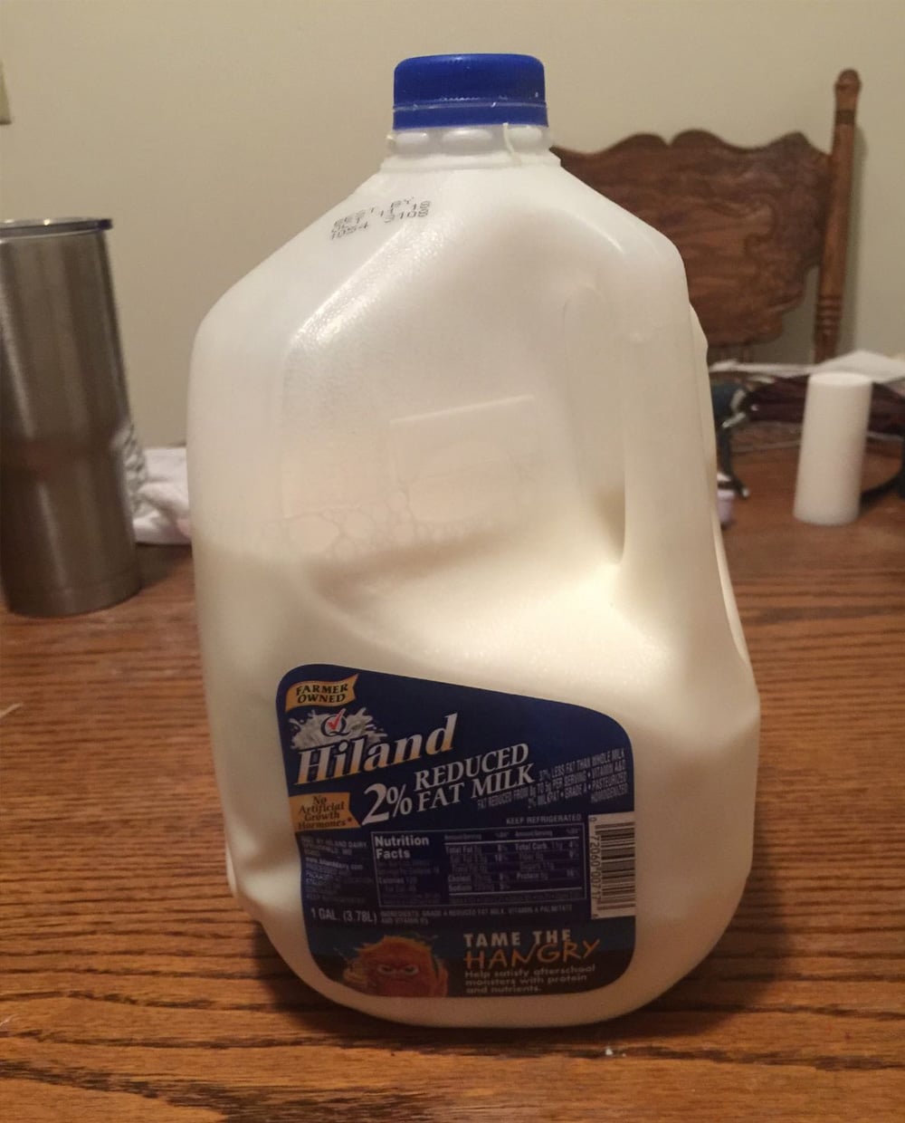 A plastic gallon jug of Hiland 2% reduced fat milk sits on a wooden table. The blue label and cap are visible, along with some household items in the background.