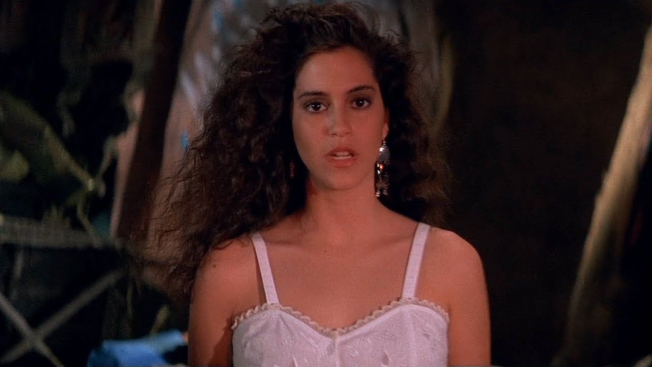 A young woman with long, wavy dark hair stands indoors, wearing a white sleeveless dress and a dangling earring, looking directly at the camera with a serious expression.