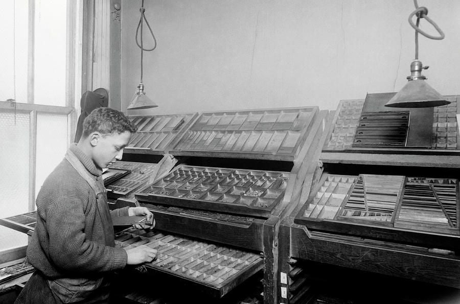 A young man sorts metal type pieces in a print shop, standing beside angled wooden type cases under hanging lamps in a room with a window and plain walls.