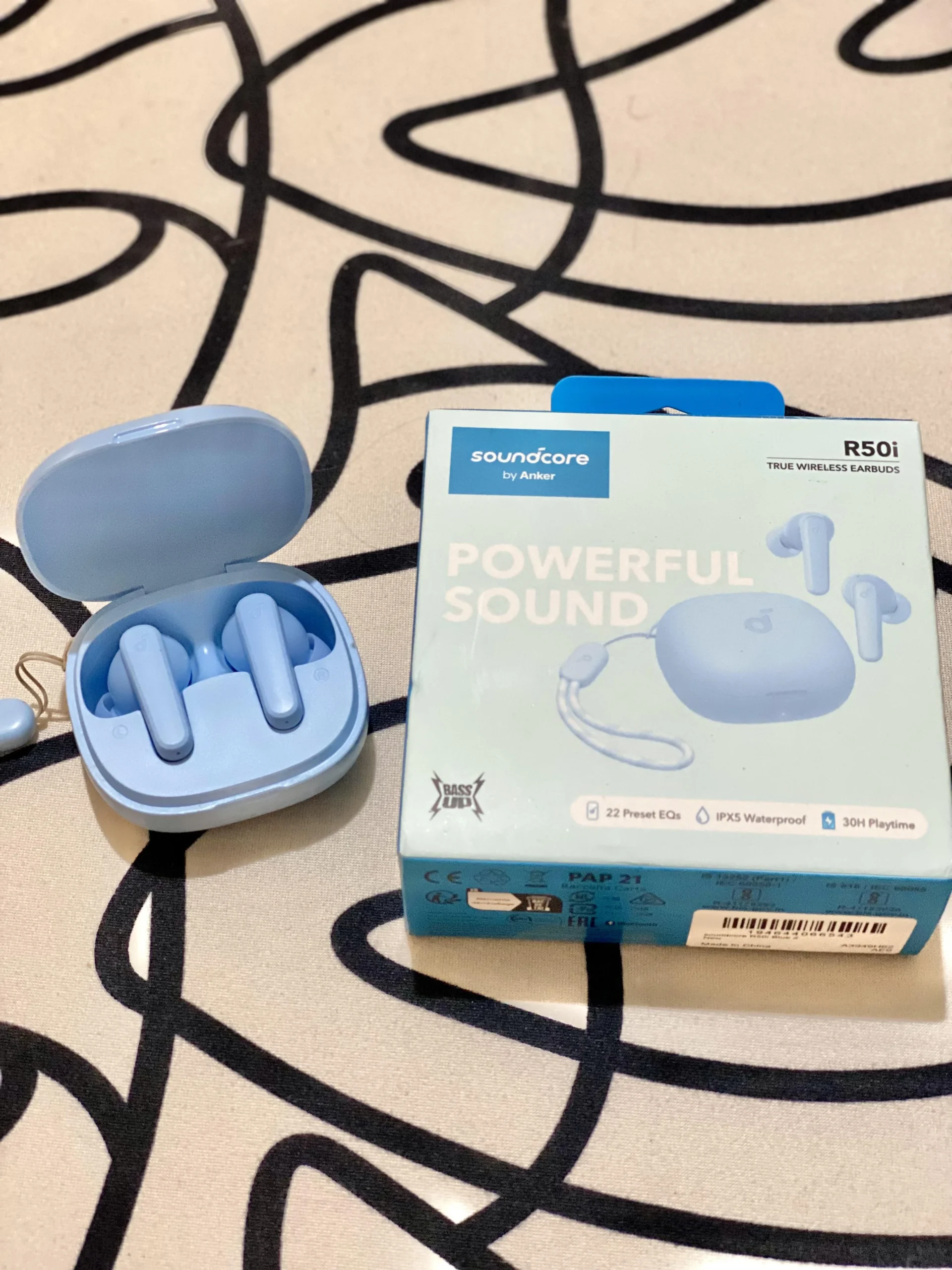 A pair of light blue wireless earbuds sit in an open charging case next to their box, which reads "Soundcore by Anker R50i Powerful Sound" with images and product features on a patterned surface.
