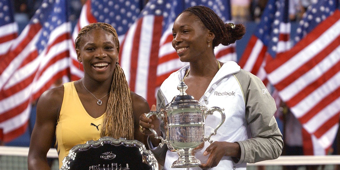 Two women tennis players smile and hold trophies on a court, with many American flags in the background. One wears yellow and holds a silver plate, the other wears a white jacket and holds a large silver cup.