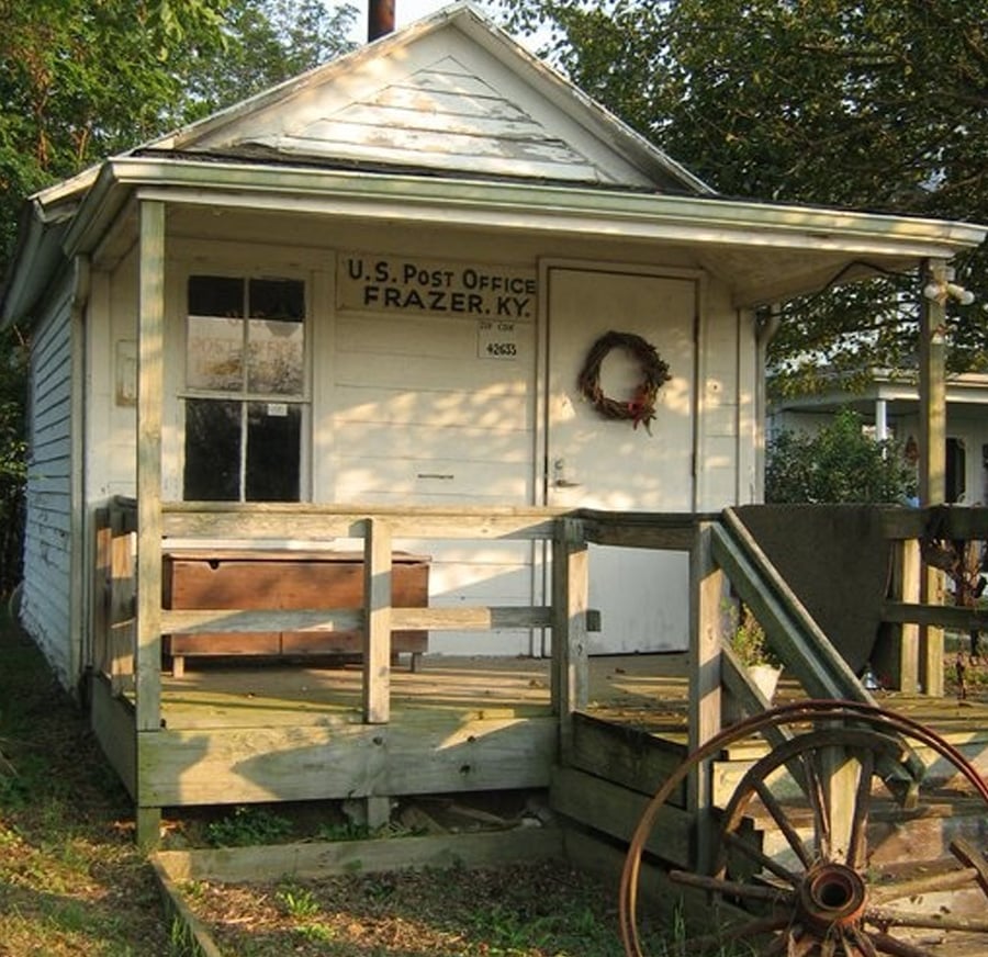 A small, old white wooden building with a porch and ramp, marked "U.S. Post Office, Frazer, KY," sits in the sunlight. A wagon wheel decorates the porch, and a wreath hangs on the door. Trees are in the background.