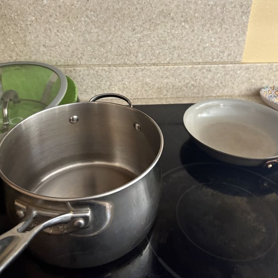 A stainless steel pot with a handle sits on a black electric stovetop next to a gray frying pan. A green-lidded pot and a dish with a colorful pattern are in the background on a beige countertop.