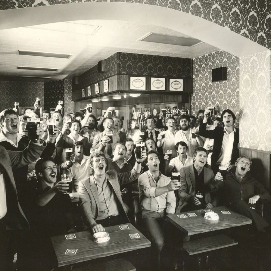 A large group of people in a vintage pub cheer and raise glasses of beer together, smiling and celebrating, surrounded by patterned wallpaper and wooden tables.