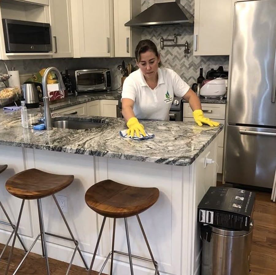 A woman wearing yellow gloves cleans a marble kitchen island with a cloth. The kitchen has white cabinets, stainless steel appliances, and two wooden stools at the counter.