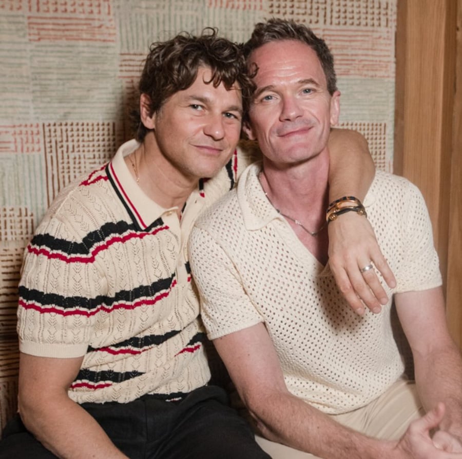 Two men sit closely together, one with his arm around the other. Both are wearing short-sleeve knit shirts and smiling gently at the camera, in a cozy indoor setting with a patterned wall behind them.