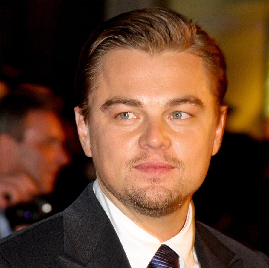 A man with light skin, short slicked-back hair, and light facial hair is wearing a dark suit, white shirt, and dark tie, looking slightly to the side at an event with blurred people in the background.