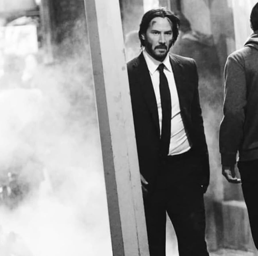 A man in a suit with long hair and a beard stands partially behind a pillar, looking serious. Smoke or mist surrounds the scene, and another person is walking away with their back to the camera. The image is in black and white.