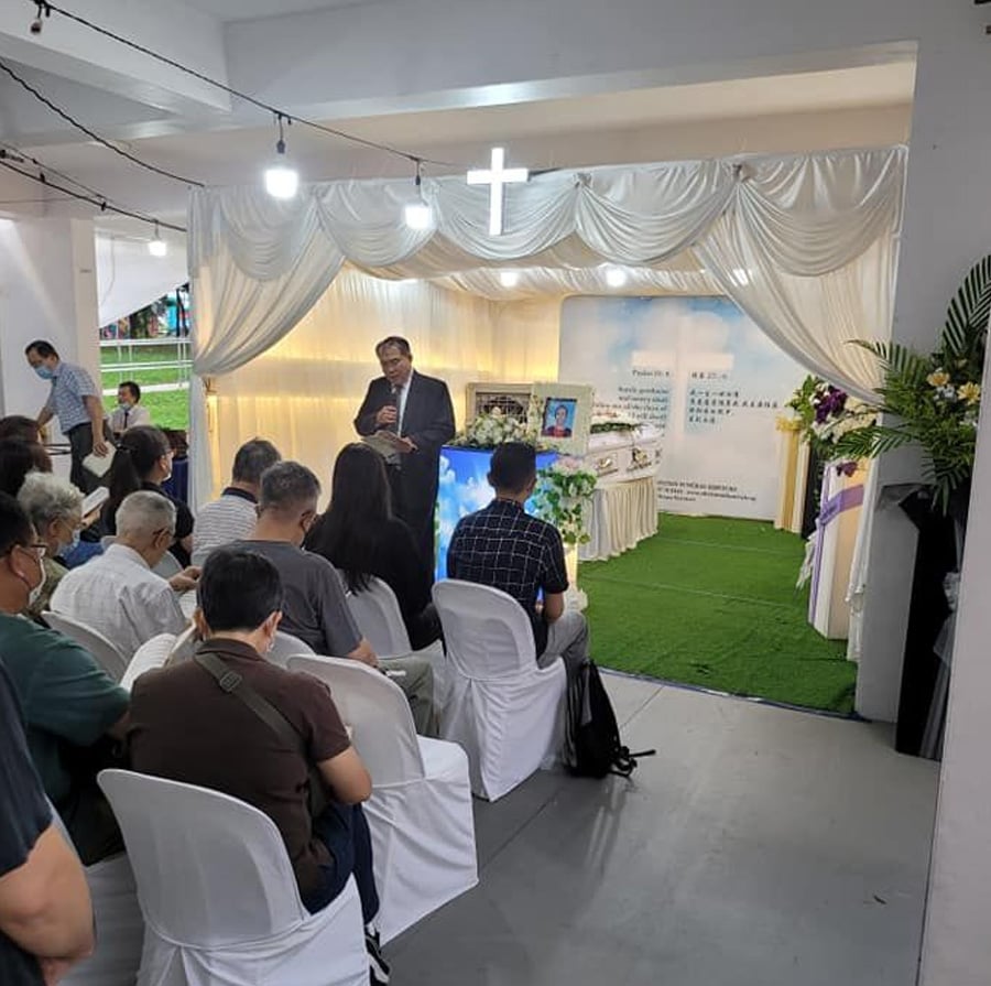 A group of people seated indoors facing a man speaking at a memorial service; white drapery, a cross, flowers, and photographs decorate the area at the front.