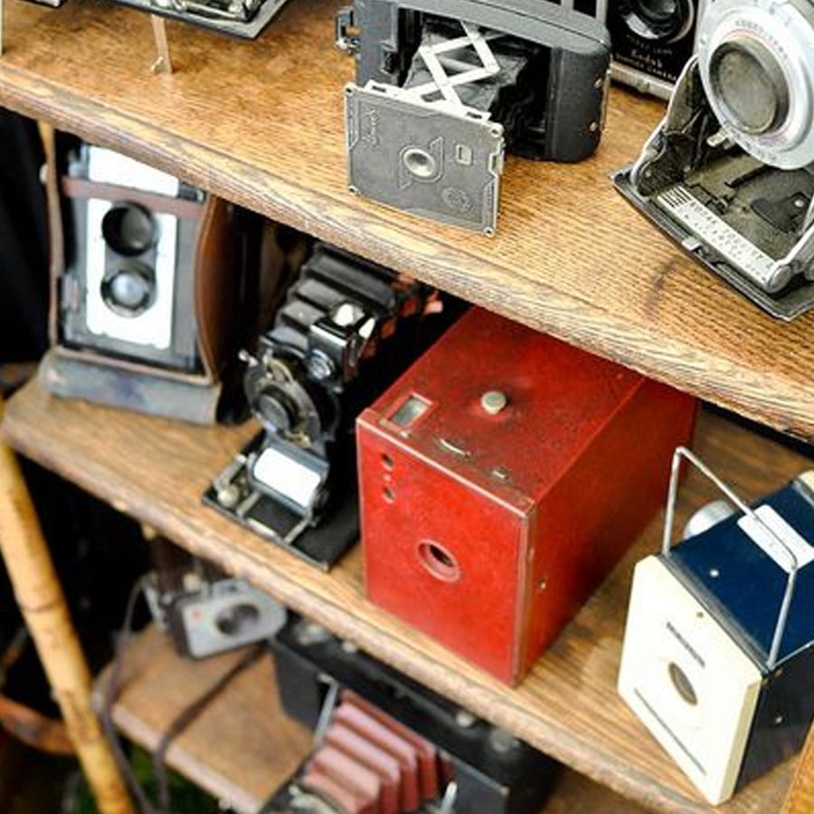 A wooden shelf displays various vintage cameras, including box cameras, folding cameras, and a large red camera, each with unique designs and different shapes.