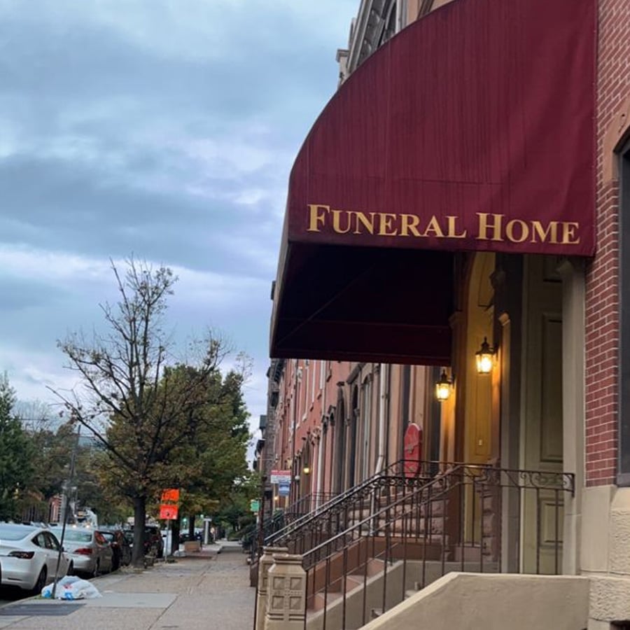 A city street with parked cars and trees runs beside a brick building with a red awning labeled "Funeral Home" in gold letters. The building has lit lamps by the entrance and a cloudy sky overhead.