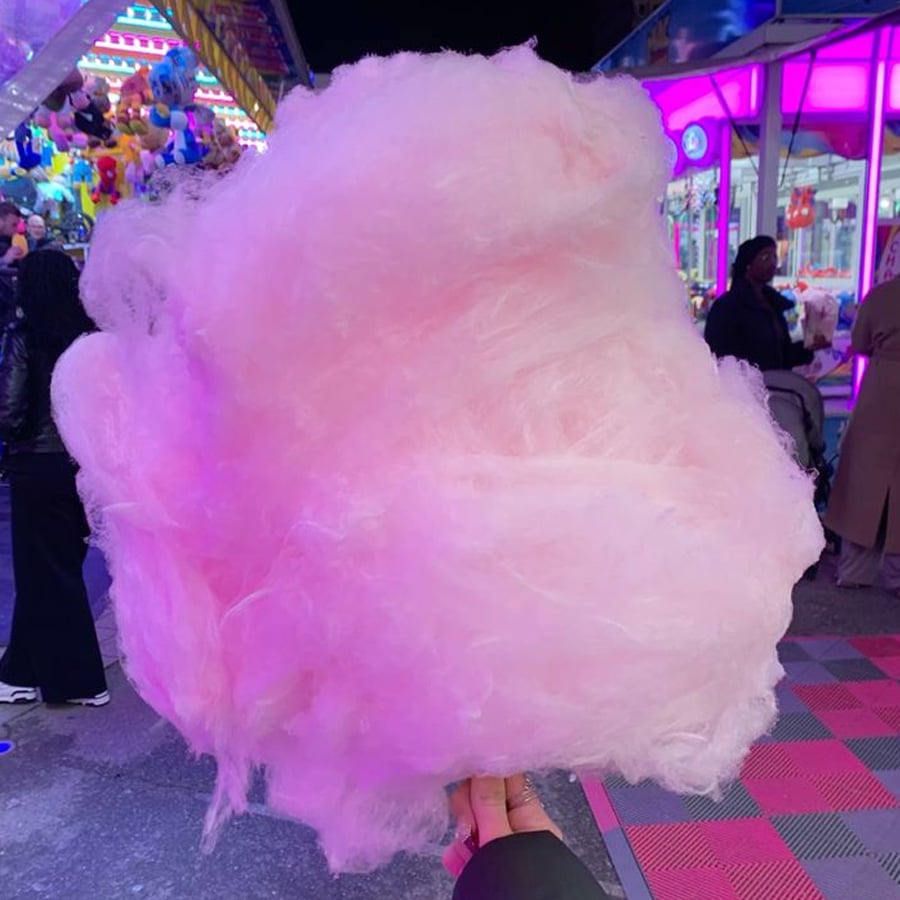 A large, fluffy pink cotton candy is held up at a colorful nighttime fair, with bright lights, game booths, and people in the background.