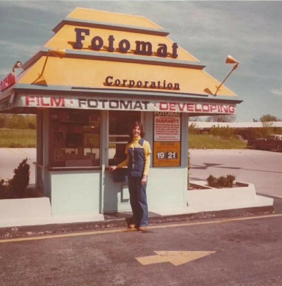 A woman in overalls stands in front of a small yellow Fotomat kiosk advertising film developing services. The kiosk has a distinctive peaked roof and is set in a parking lot on a sunny day.