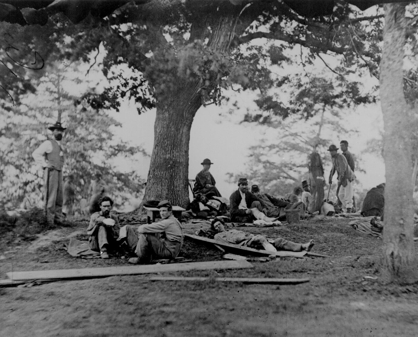 A group of soldiers rest and sit under large trees, some lying on the ground, during the Civil War era. The scene is outdoors with scattered boards and a somber atmosphere.
