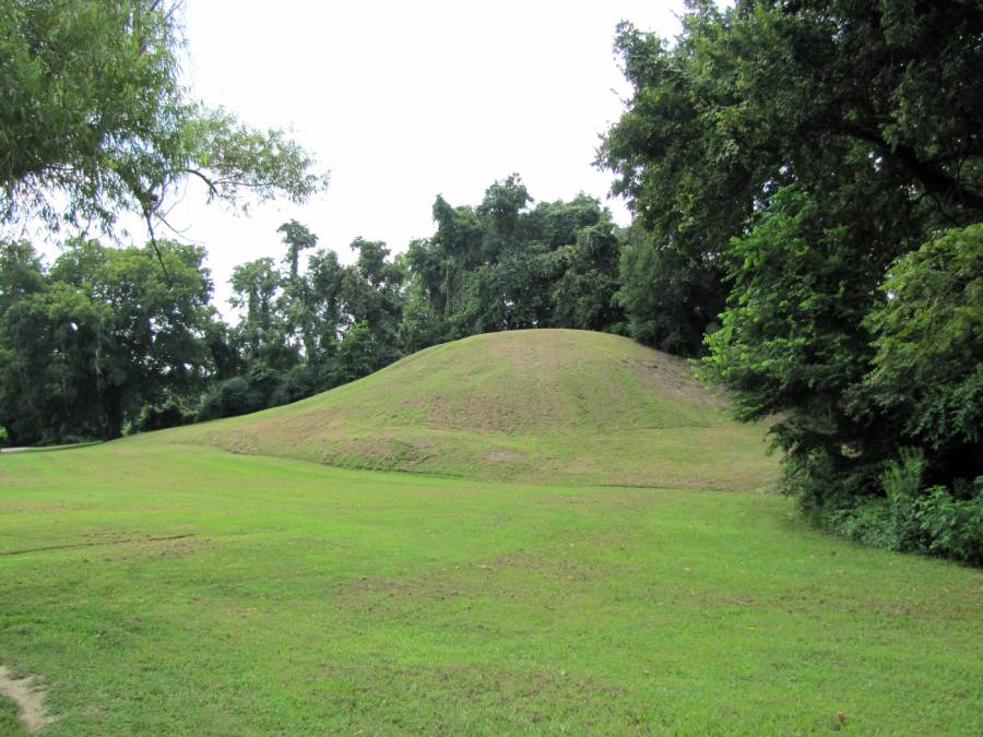 A grassy earthen mound rises gently from a well-maintained lawn, surrounded by dense green trees under a cloudy sky.