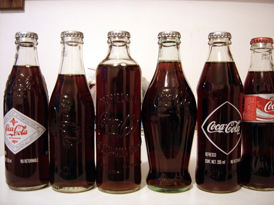 Six vintage Coca-Cola glass bottles with different labels and shapes are displayed side by side on a white surface against a plain background.