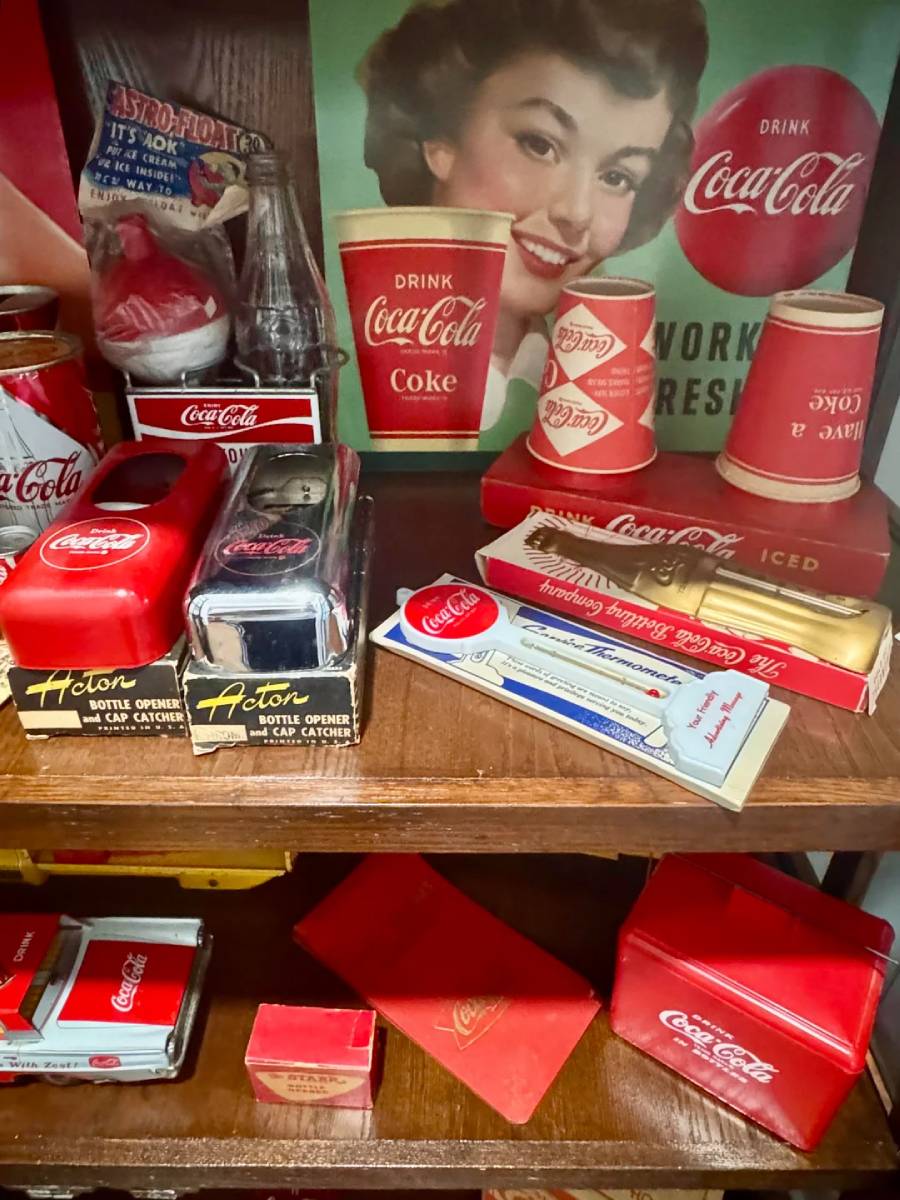 A display shelf with vintage Coca-Cola memorabilia, including red cups, toy cars, bottle openers, and advertisements, with a smiling woman in a retro Coca-Cola poster in the background.