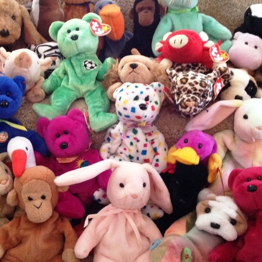 A collection of colorful Beanie Babies, including animals like a pink bunny, green bear, purple duck, and leopard, are arranged closely together on a carpeted floor. Each has a Ty tag attached to its ear.