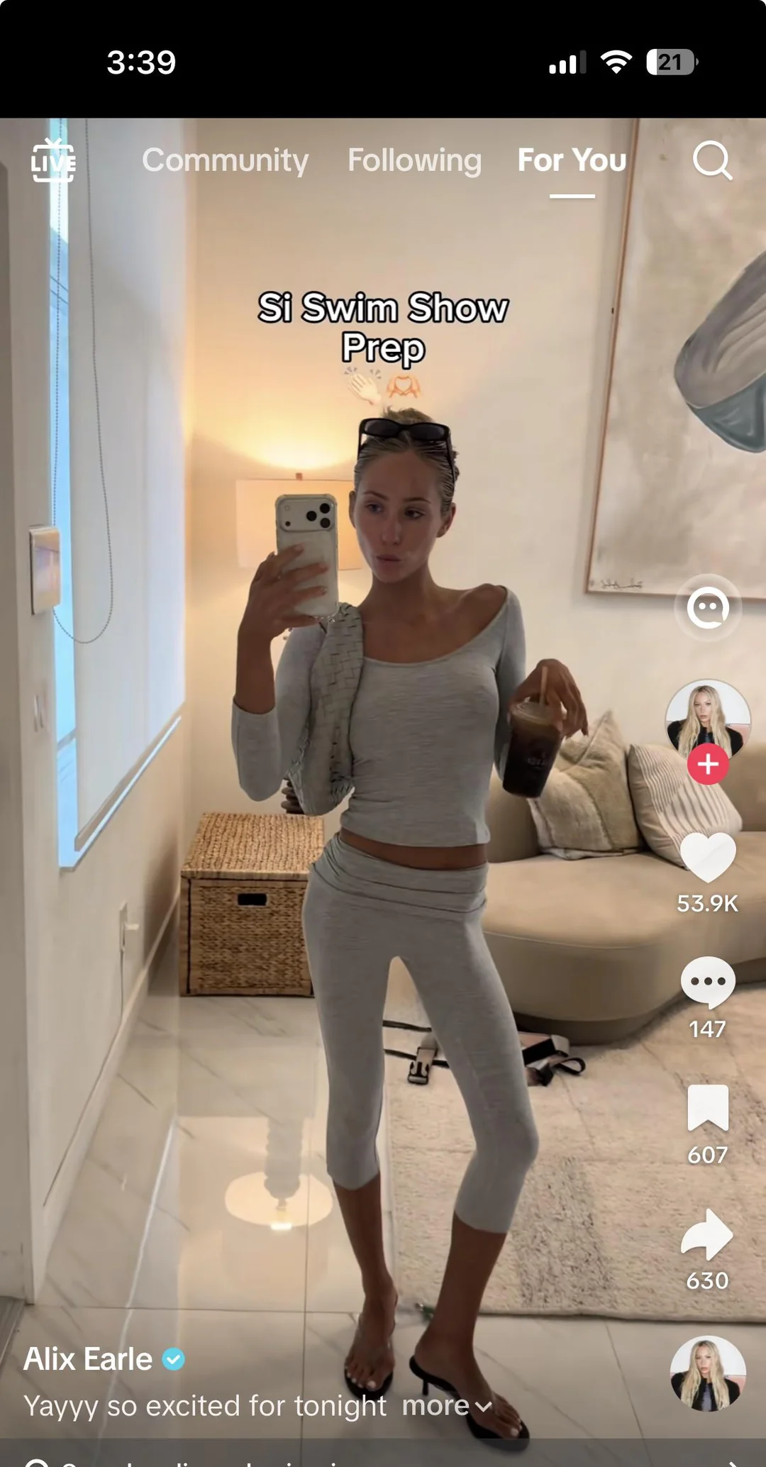 A woman in a light gray matching set takes a mirror selfie while holding a beverage and a bag. The screen shows TikTok interface elements, with the caption “Si Swim Show Prep” and username “Alix Earle.”
