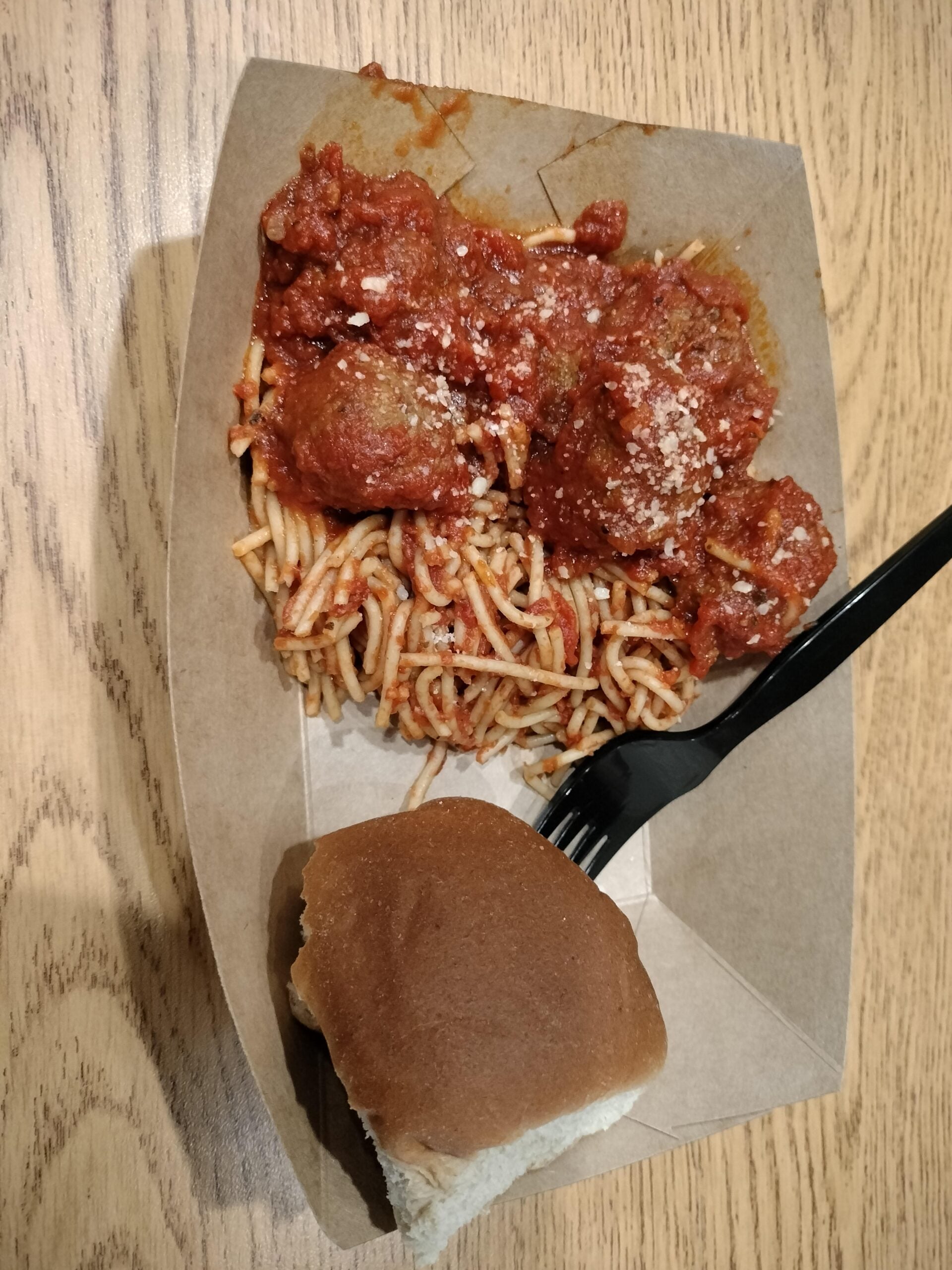A cardboard tray containing spaghetti with tomato sauce and meatballs, topped with grated cheese, a plastic fork, and a brown bread roll on a wooden table.