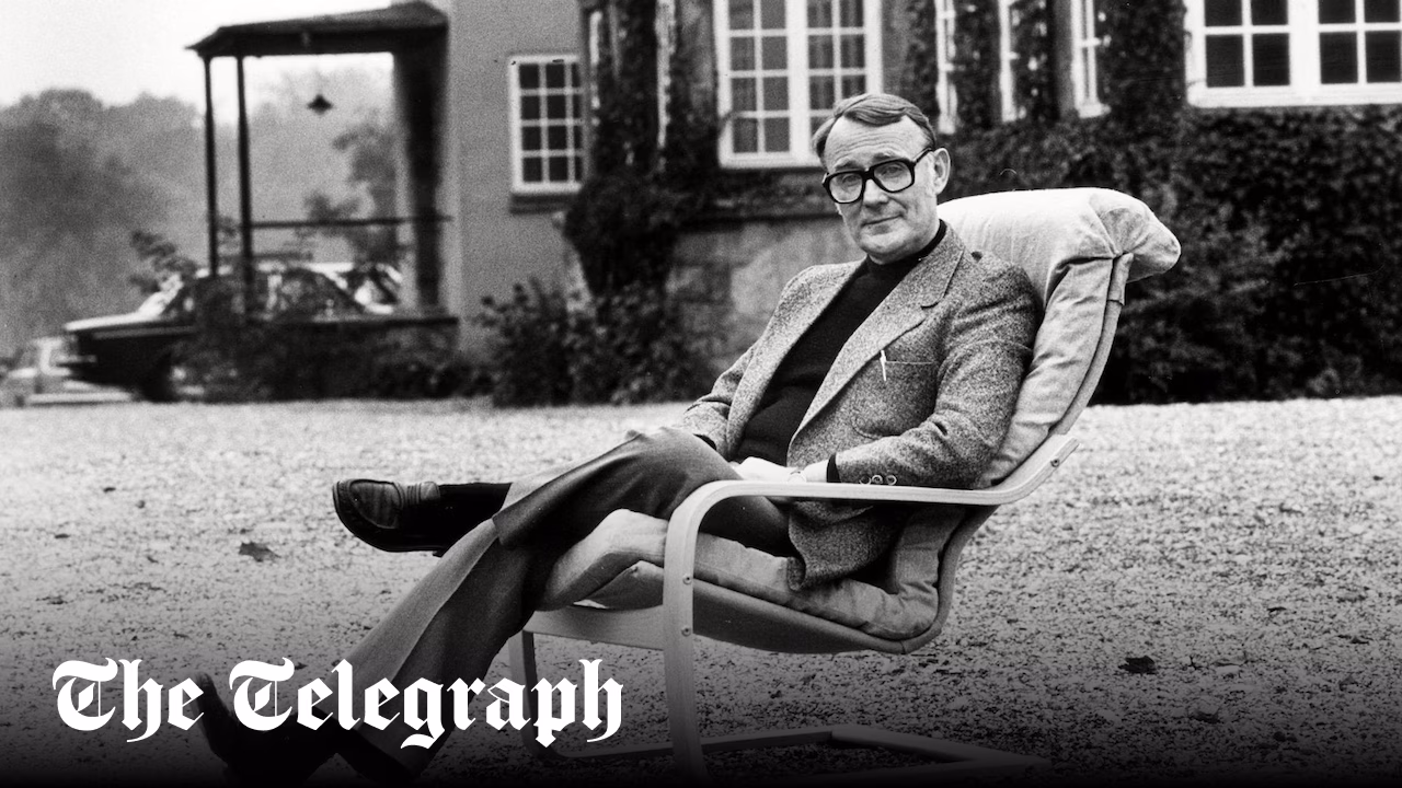 A man in glasses and a suit jacket sits in a reclining chair outdoors on a gravel surface, with a building and vines in the background. The Telegraph logo appears in the lower left corner.